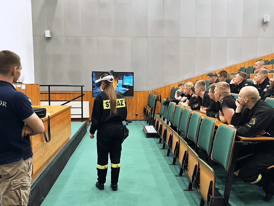 Ein Feuerwehranwärter mit VR-Brille vor einem Hörsaal voller Feuerwehrleute, die VR-Ansicht ist auf einer Leinwand zu sehen