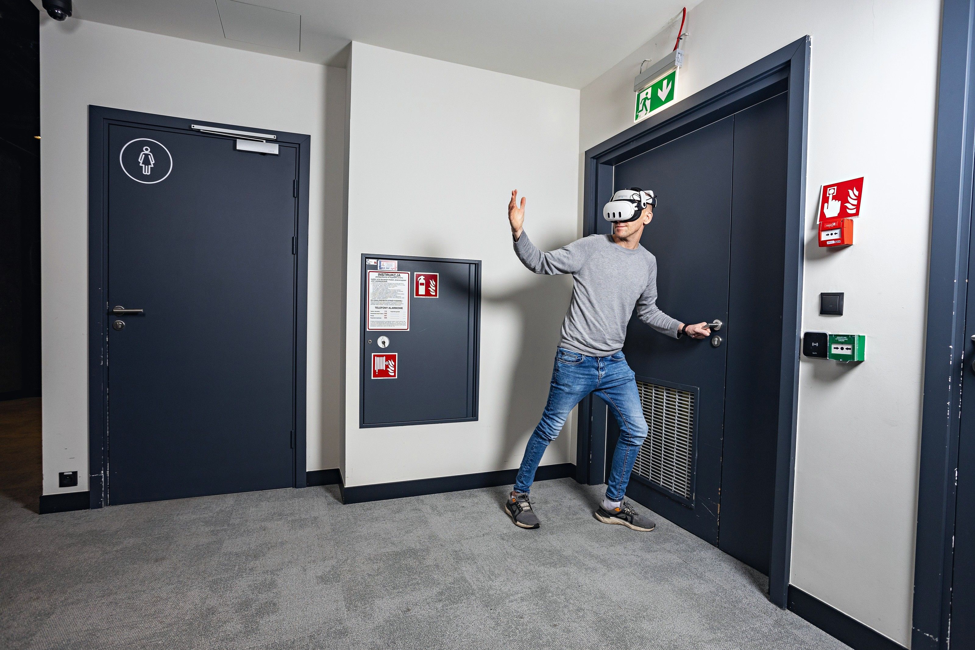 An employee in a VR headset practises opening a door in an office corridor next to fire safety signs