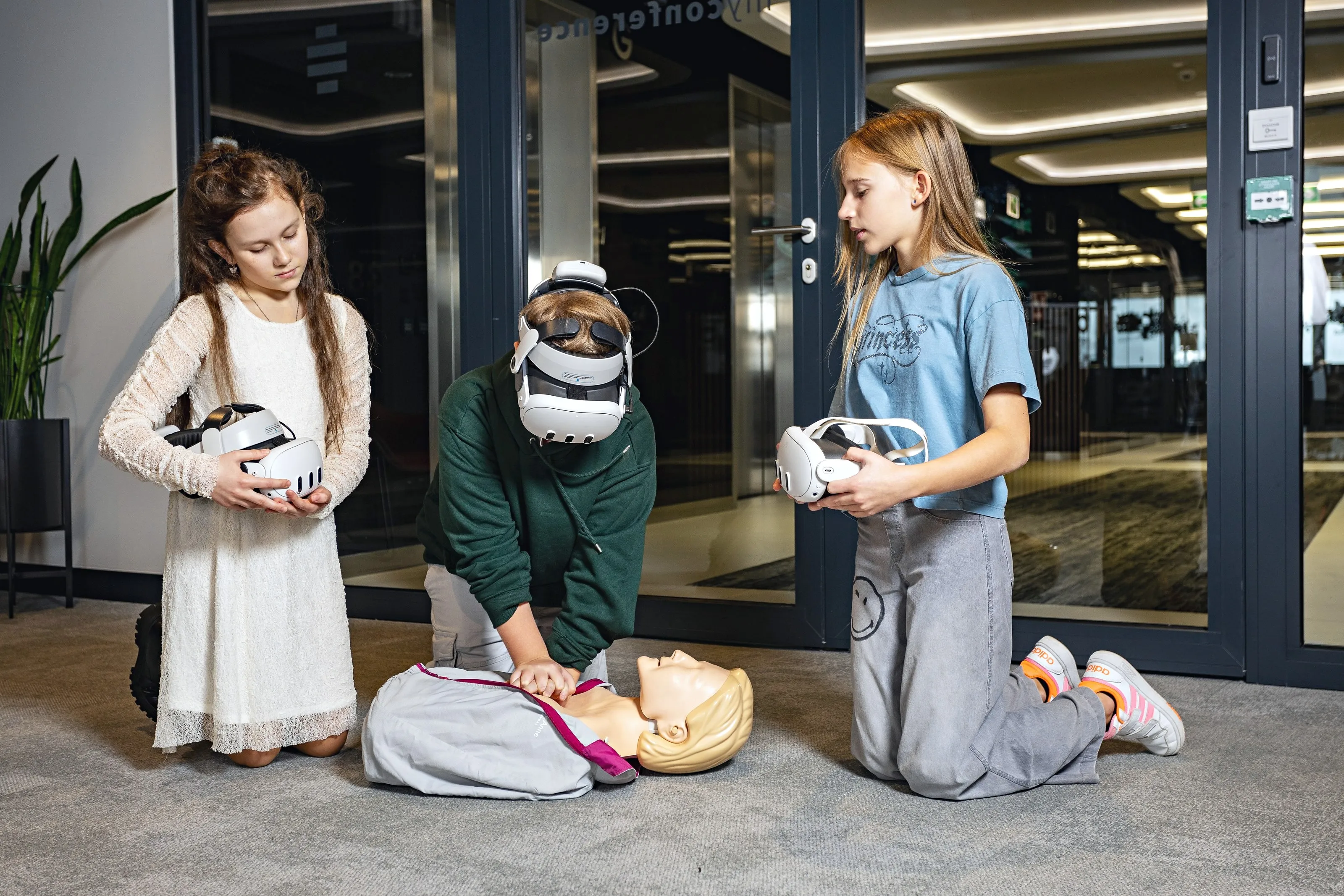 A boy in a VR headset practises CPR on a mannequin while two girls hold their headsets